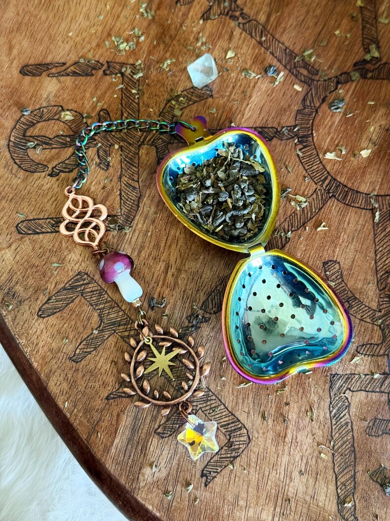 May include: Iridescent heart-shaped tea infuser filled with loose tea. The infuser is open, revealing the tea leaves. A chain with decorative charms, including a mushroom, star, and a crystal star, hangs from the infuser. The background is a wooden surface.