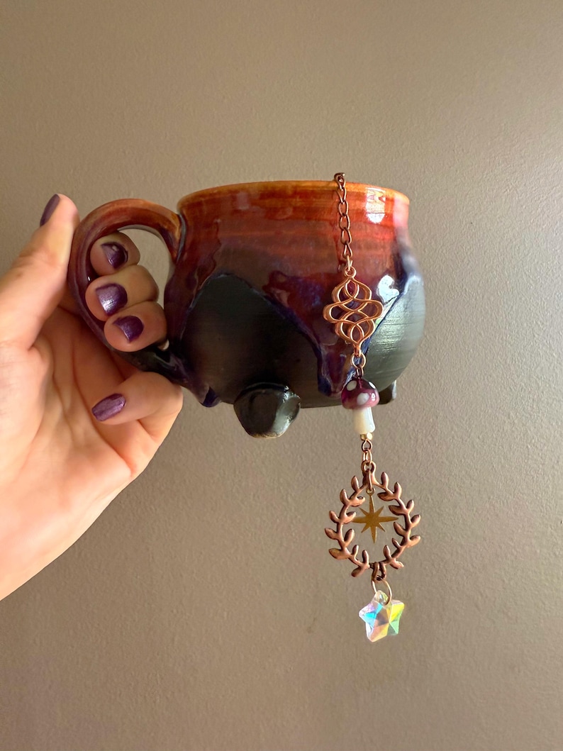 May include: A handmade ceramic mug with a dark red, orange, and black glaze. A copper charm with a star and crystal hangs from the rim. The mug has a unique shape and a sturdy handle, perfect for enjoying a warm beverage.