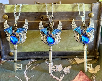 Puede incluir: Tres collares de llave plateados con alas de dragón azules y una piedra preciosa azul. El dragón está enroscado alrededor de la llave.