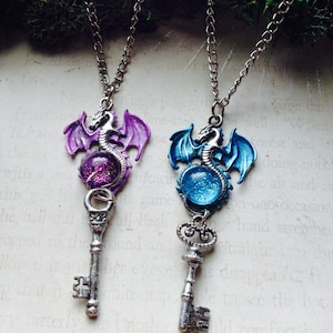 May include: Two fantasy-themed necklaces with silver chains. Each pendant features a dragon atop a key. One dragon is purple, the other blue, both with a glittery orb. The keys are silver-toned.