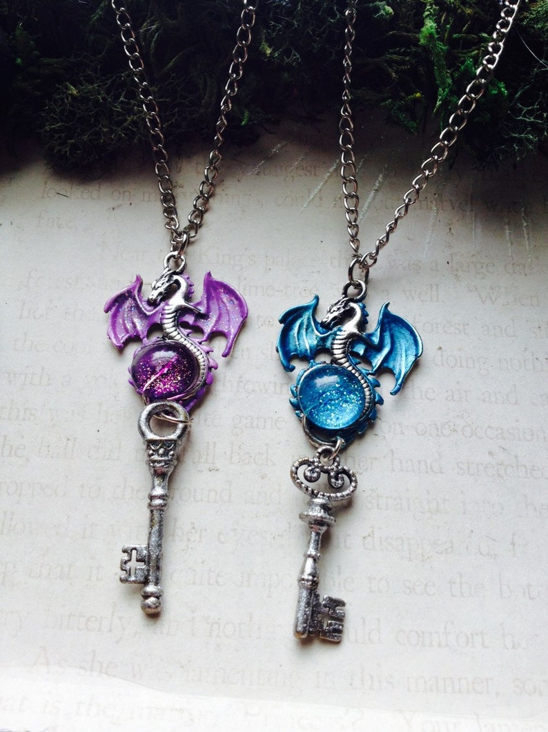 May include: Two fantasy-themed necklaces with silver chains. Each pendant features a dragon atop a key. One dragon is purple, the other blue, both with a glittery orb. The keys are silver-toned.
