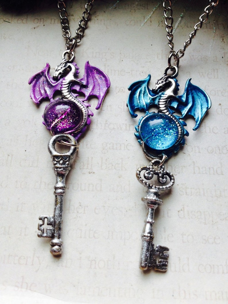 May include: Two fantasy-themed necklaces. Each features a silver-colored key pendant topped with a dragon and a glitter-filled orb. One dragon is purple, the other blue, both with matching wings. The keys hang from silver chains.