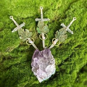 Op de afbeelding: Drie decoratieve zwaardvormige bedels met bladeren en een centrale amethistkristal. De zwaarden zijn zilverkleurig en de bladeren hebben een groen-gouden afwerking. De amethist is een mix van paarse en witte tinten.