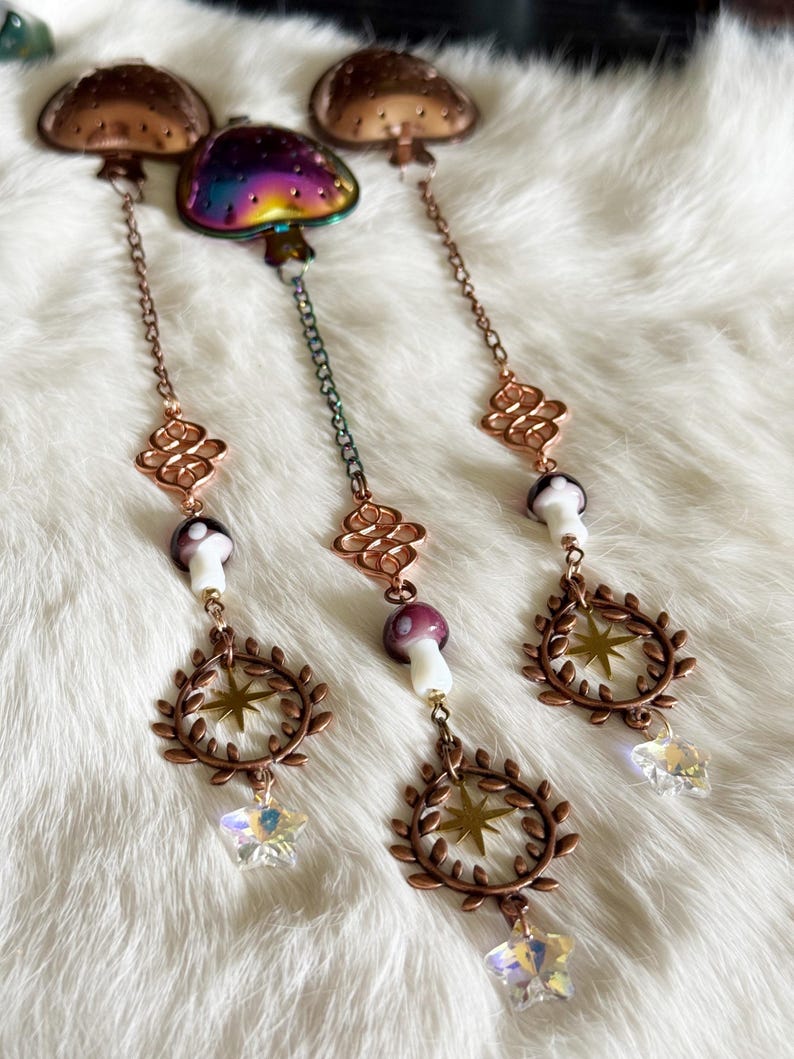May include: Three mushroom-themed hanging ornaments. The top mushrooms are bronze-colored, with a rainbow-colored mushroom in the center. Each ornament has a chain, a decorative element, a mushroom, a star, a wreath, and a crystal star.