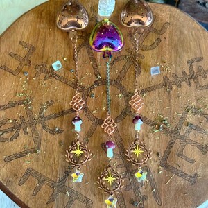 May include: A collection of decorative tea infusers and charms. The infusers are heart-shaped, with a copper finish, and hang from chains. The charms include mushrooms, starbursts, and crystal stars. The items are displayed on a wooden surface with etched designs.