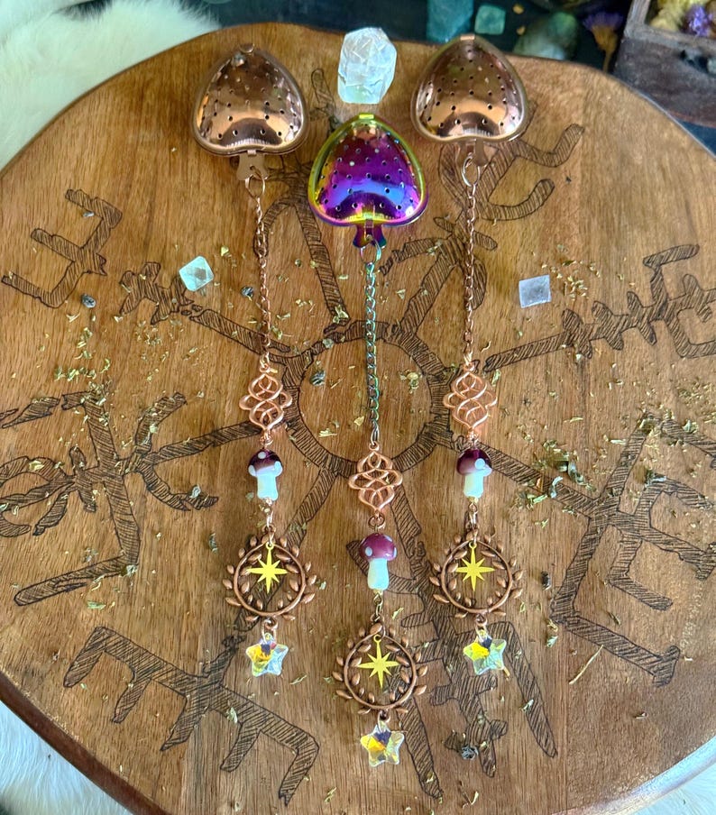 May include: A collection of decorative tea infusers and charms. The infusers are heart-shaped, with a copper finish, and hang from chains. The charms include mushrooms, starbursts, and crystal stars. The items are displayed on a wooden surface with etched designs.