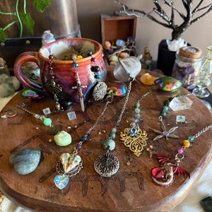 May include: A collection of tea infusers with various charms and crystals. The infusers are made of metal and have different designs, including a dragon, a dragonfly, a heart, and a flower. The charms are made of metal and glass and are in various colors, including gold, silver, red, green, and blue. The crystals are clear and have a variety of shapes and sizes.