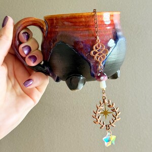 May include: A handmade ceramic mug with a unique glaze in shades of red, purple, and black. The mug features a decorative copper tea infuser with a mushroom, wreath, and star charm. The mug has three small feet.