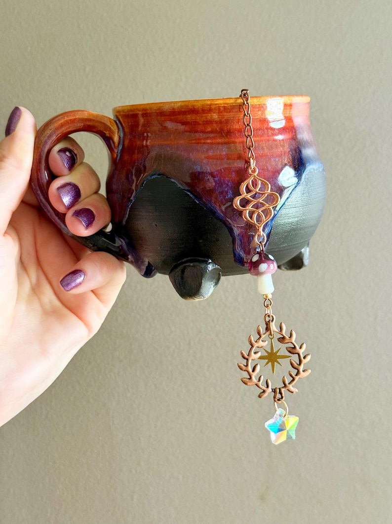 May include: A handmade ceramic mug with a unique glaze in shades of red, purple, and black. The mug features a decorative copper tea infuser with a mushroom, wreath, and star charm. The mug has three small feet.