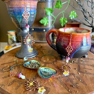 May include: A ceramic mug and goblet with a red, orange, and blue glaze, alongside a wooden tea tray. The tray holds two tea infusers and decorative copper charms with star and mushroom designs. The scene evokes a whimsical tea party.