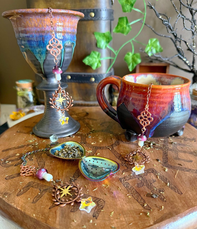 May include: A ceramic mug and goblet with a red, orange, and blue glaze, alongside a wooden tea tray. The tray holds two tea infusers and decorative copper charms with star and mushroom designs. The scene evokes a whimsical tea party.
