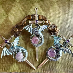 May include: Three fantasy-themed hair accessories. Each features a silver dragon with blue wings, a purple gemstone, and a sword design. The accessories are set against a textured, olive-green background.
