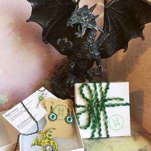 May include: A black dragon statue with outstretched wings is the centerpiece of this image. Jewelry, including a dragon pendant necklace and earrings with eye designs, is displayed in white boxes. A gift box is tied with green yarn.