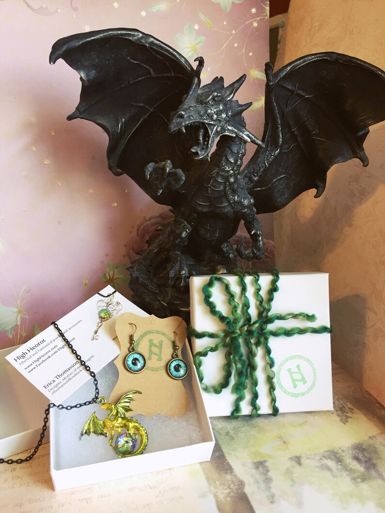 May include: A black dragon statue with outstretched wings is the centerpiece of this image. Jewelry, including a dragon pendant necklace and earrings with eye designs, is displayed in white boxes. A gift box is tied with green yarn.