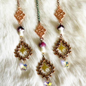 May include: Three hanging decorative charms. Each charm features a copper-colored filigree, a mushroom with a white stem and purple cap, a wreath with a yellow star, and a crystal star pendant. The charms hang from delicate chains.