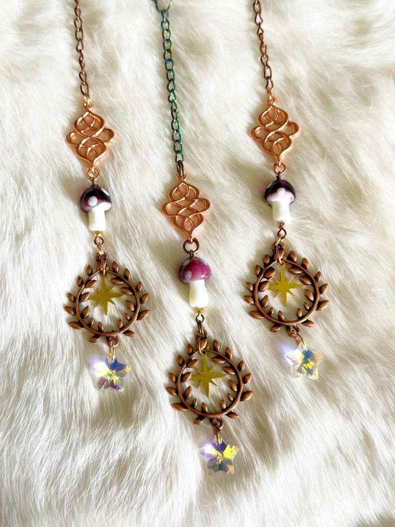 May include: Three hanging decorative charms. Each charm features a copper-colored filigree, a mushroom with a white stem and purple cap, a wreath with a yellow star, and a crystal star pendant. The charms hang from delicate chains.