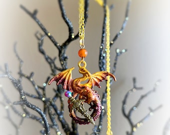 May include: A gold chain necklace with a red and orange dragon pendant holding a sword. The dragon has a purple gem on its head and is set against a black branch background.