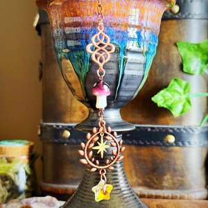 May include: A copper-colored charm hangs in front of a ceramic vase. The charm features an infinity symbol, a mushroom, a wreath with a star, and a yellow star. The vase is brown, blue, and orange.