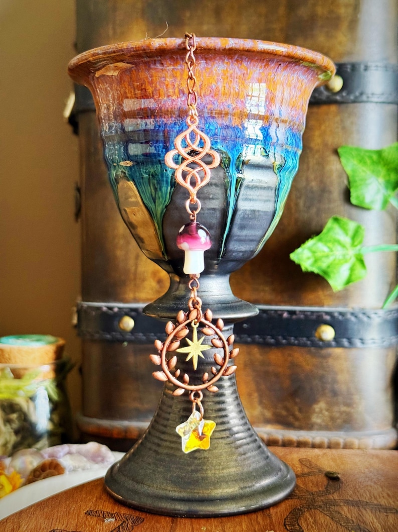 May include: A copper-colored charm hangs in front of a ceramic vase. The charm features an infinity symbol, a mushroom, a wreath with a star, and a yellow star. The vase is brown, blue, and orange.