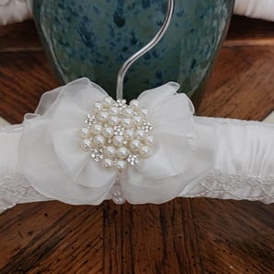 May include: A white satin padded hanger with a decorative floral embellishment. The embellishment features a cluster of faux pearls and sparkling rhinestones, set against a backdrop of layered white fabric. The hanger has a lace trim.