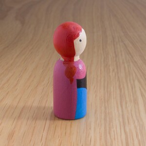 May include: A painted wooden peg doll with a red head and hair, a white face, and a pink body. The doll has a black and blue section on the side. The doll is standing on a wooden surface.