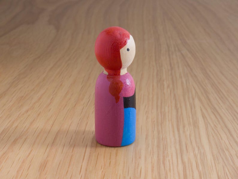 May include: A painted wooden peg doll with a red head and hair, a white face, and a pink body. The doll has a black and blue section on the side. The doll is standing on a wooden surface.