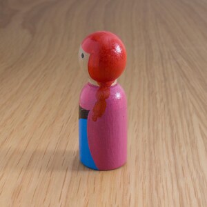 May include: A wooden peg doll with a red head and a pink dress. The doll has a blue skirt and a black stripe. The doll's red hair is styled in a braid. The doll is standing on a wooden surface.
