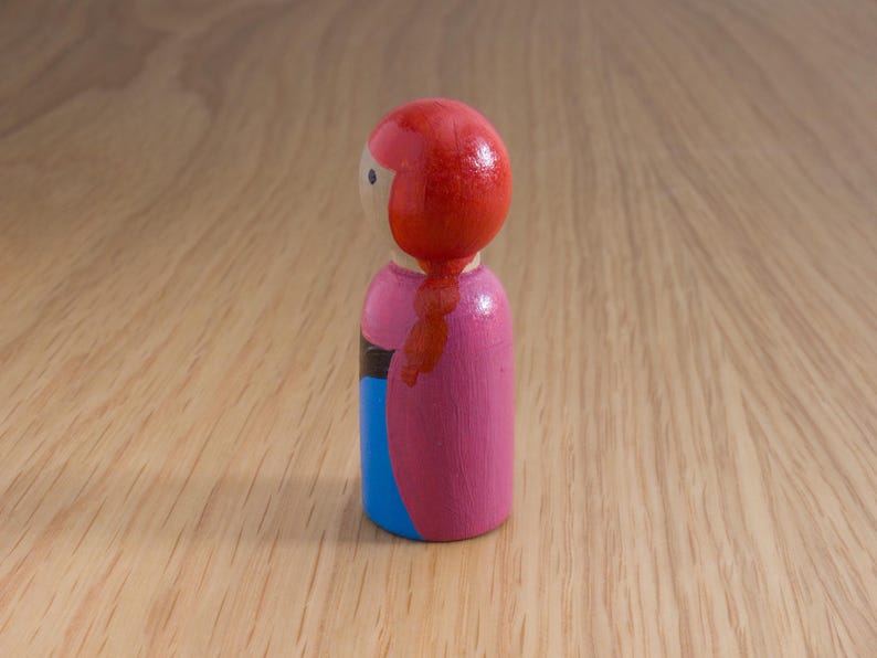May include: A wooden peg doll with a red head and a pink dress. The doll has a blue skirt and a black stripe. The doll's red hair is styled in a braid. The doll is standing on a wooden surface.