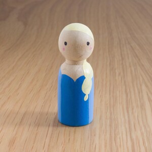May include: A wooden peg doll featuring a blue dress and blonde hair. The doll has a light skin tone, with painted facial features including small black eyes and pink cheeks. The doll is standing on a light brown wooden surface.