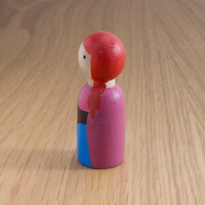 May include: A wooden peg doll with a red head and a pink body. The doll has a blue section on the bottom and a black stripe. The doll is on a wooden surface.