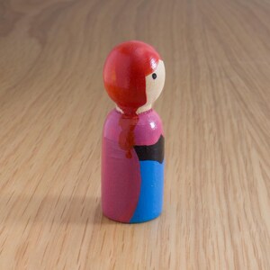 May include: A hand-painted wooden peg doll with a red head, a light-colored face, and a pink body with black and blue accents. The doll is standing on a light-colored wooden surface.