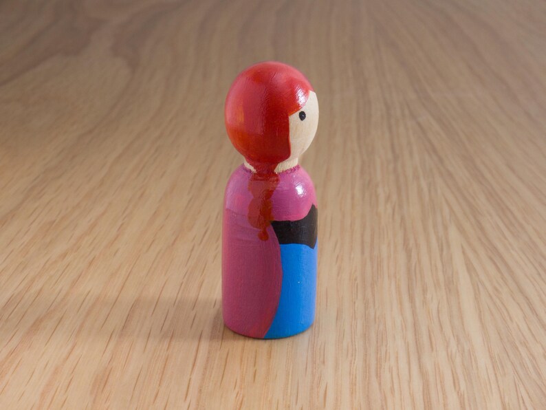 May include: A hand-painted wooden peg doll with a red head, a light-colored face, and a pink body with black and blue accents. The doll is standing on a light-colored wooden surface.