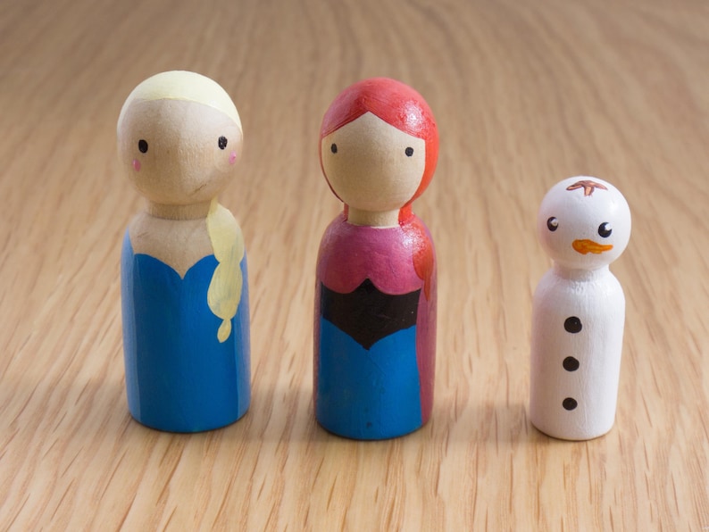 May include: Three wooden peg dolls: two female figures and a snowman. The figures have painted details, including blue, pink, and red clothing. The snowman is white with black button eyes and a carrot nose. The dolls are on a wooden surface.