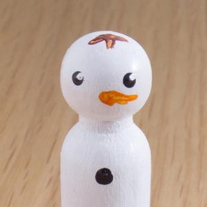 May include: A handcrafted snowman peg doll with a white body, black eyes, and a carrot-orange nose. A small brown design is painted on the top of the head, and a black dot is on the body. This decorative item is perfect for winter holiday decor.