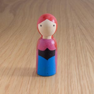 May include: A wooden peg doll with a red head and hair, a pink top, a black bodice, and a blue skirt. The doll has a simple, painted face and stands on a wooden surface.