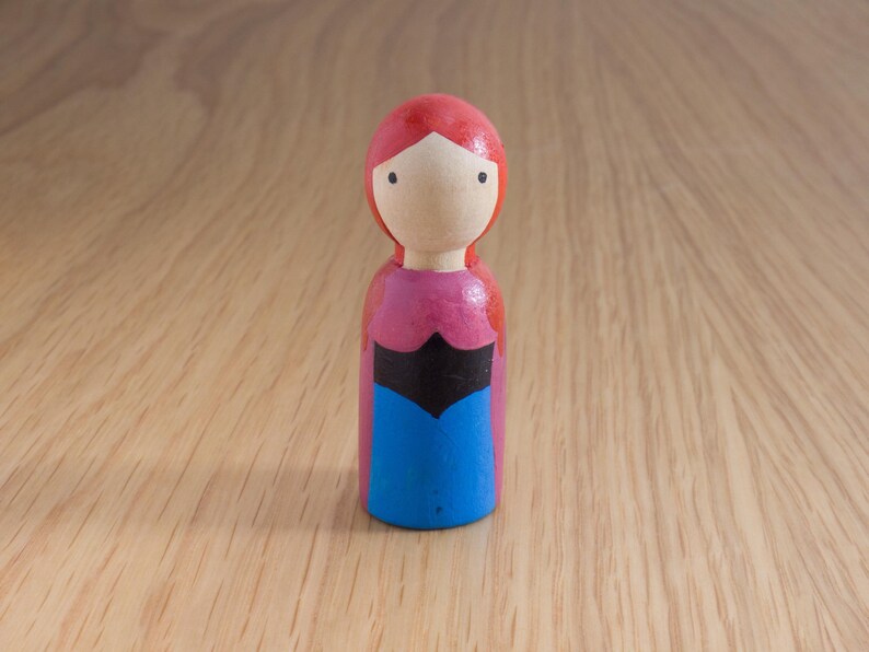 May include: A wooden peg doll with a red head and hair, a pink top, a black bodice, and a blue skirt. The doll has a simple, painted face and stands on a wooden surface.