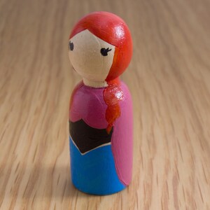 May include: A hand-painted wooden peg doll featuring a character with red hair in a side braid. The doll wears a pink and blue dress with a black bodice. The doll has black eyes and is set against a light wood background.
