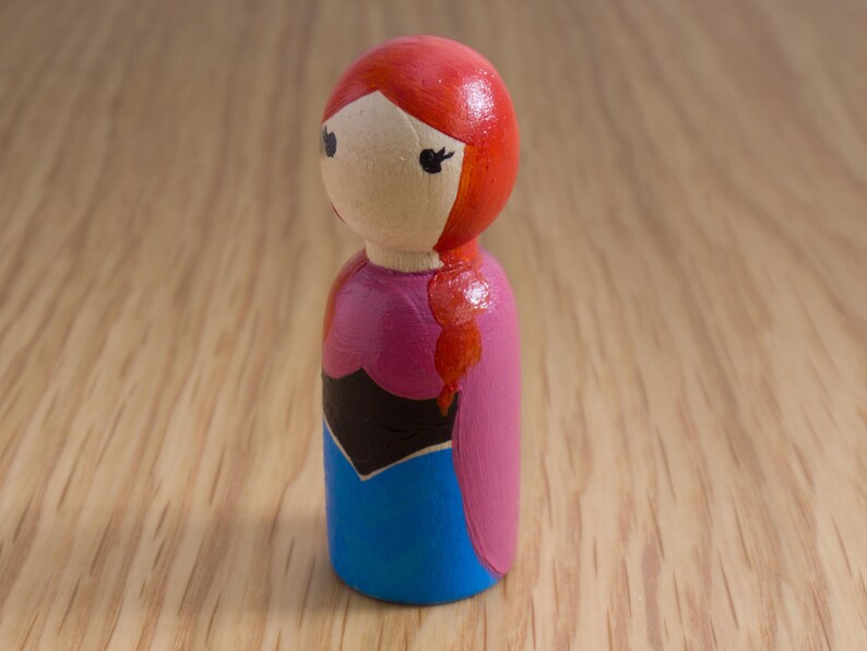 May include: A hand-painted wooden peg doll featuring a character with red hair in a side braid. The doll wears a pink and blue dress with a black bodice. The doll has black eyes and is set against a light wood background.