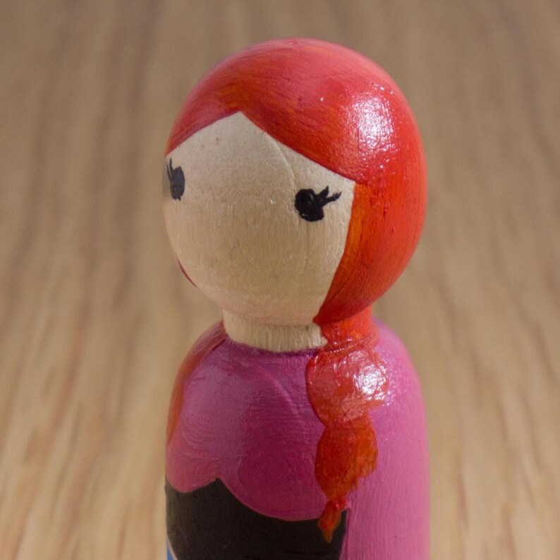 May include: A hand-painted wooden peg doll with red hair and a pink dress. The doll's face is painted with black eyes and a red mouth. The hair is styled in a side part and a braid.