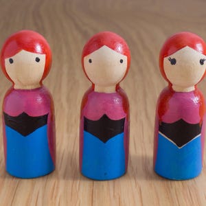 May include: Three wooden peg dolls with red hair, painted faces, and colorful clothing. The dolls have pink tops, black accents, and blue skirts. They are standing on a wooden surface.