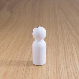 May include: A small, white wooden peg doll with a rounded head and cylindrical body. The top of the head has a small, brown painted design. The doll is standing on a light brown wooden surface.