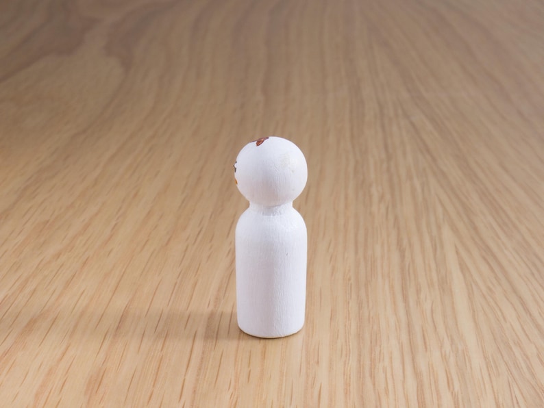 May include: A small, white wooden peg doll with a rounded head and cylindrical body. The top of the head has a small, brown painted design. The doll is standing on a light brown wooden surface.