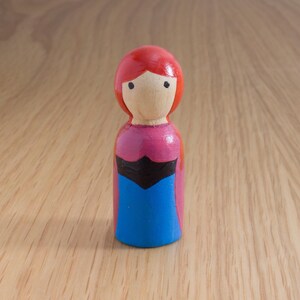 May include: A hand-painted wooden peg doll with a red head, a light wooden face, and a pink and blue dress. The doll has black accents and is standing on a wooden surface. The doll is a handmade craft.
