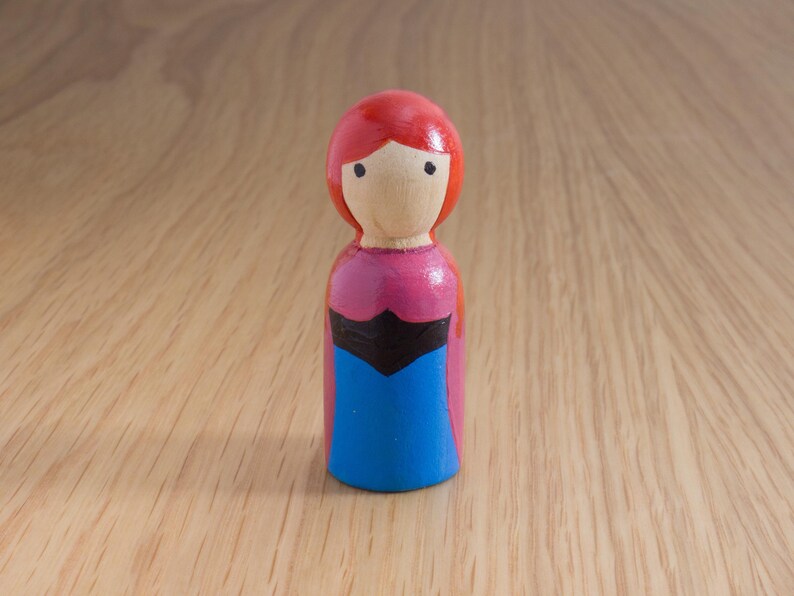 May include: A hand-painted wooden peg doll with a red head, a light wooden face, and a pink and blue dress. The doll has black accents and is standing on a wooden surface. The doll is a handmade craft.