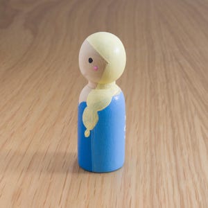 May include: A wooden peg doll featuring a painted design. The doll has a light yellow head and hair, a blue dress, and a small pink blush on the cheeks. The doll is standing on a wooden surface.