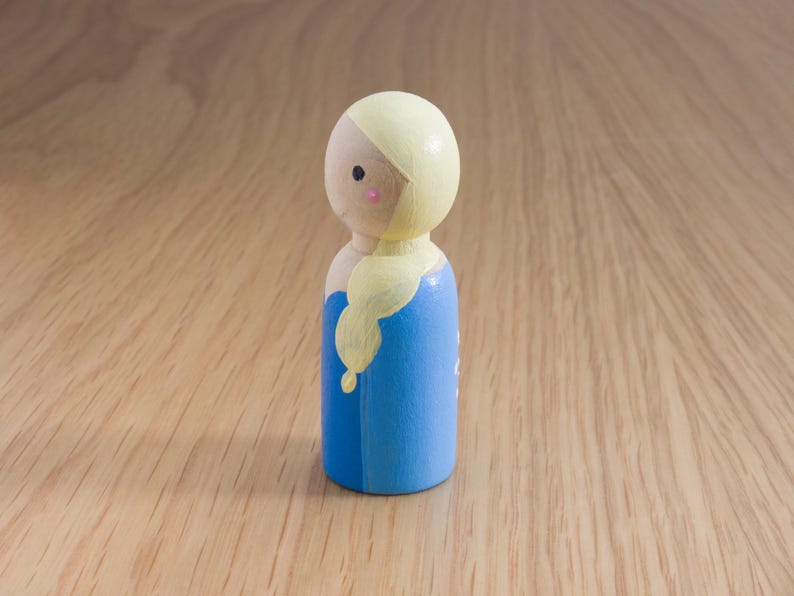 May include: A wooden peg doll featuring a painted design. The doll has a light yellow head and hair, a blue dress, and a small pink blush on the cheeks. The doll is standing on a wooden surface.