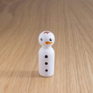May include: A small, white wooden snowman figurine with black dot buttons and a painted orange carrot nose. The snowman has black eyes and a small brown design on top of its head. The figurine is standing on a light brown wooden surface.