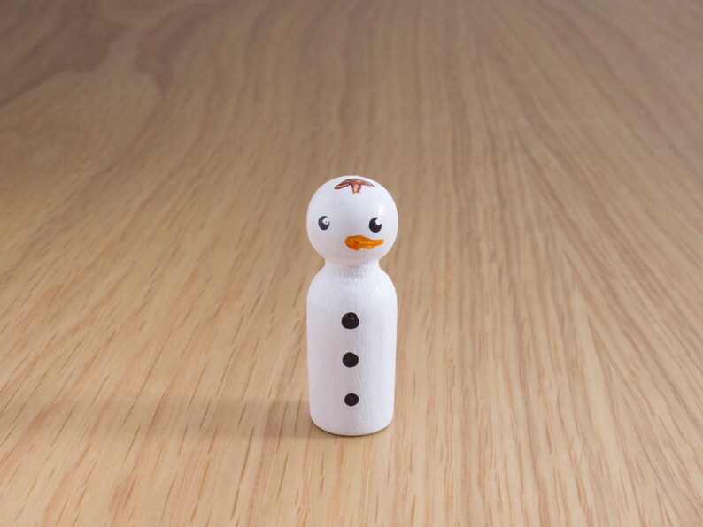May include: A small, white wooden snowman figurine with black dot buttons and a painted orange carrot nose. The snowman has black eyes and a small brown design on top of its head. The figurine is standing on a light brown wooden surface.