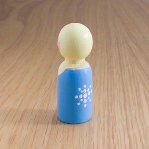 May include: A painted wooden peg doll with a light yellow head and a blue body. The blue body features a white dot design. The doll is standing on a wooden surface.