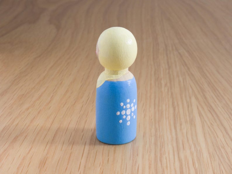 May include: A painted wooden peg doll with a light yellow head and a blue body. The blue body features a white dot design. The doll is standing on a wooden surface.